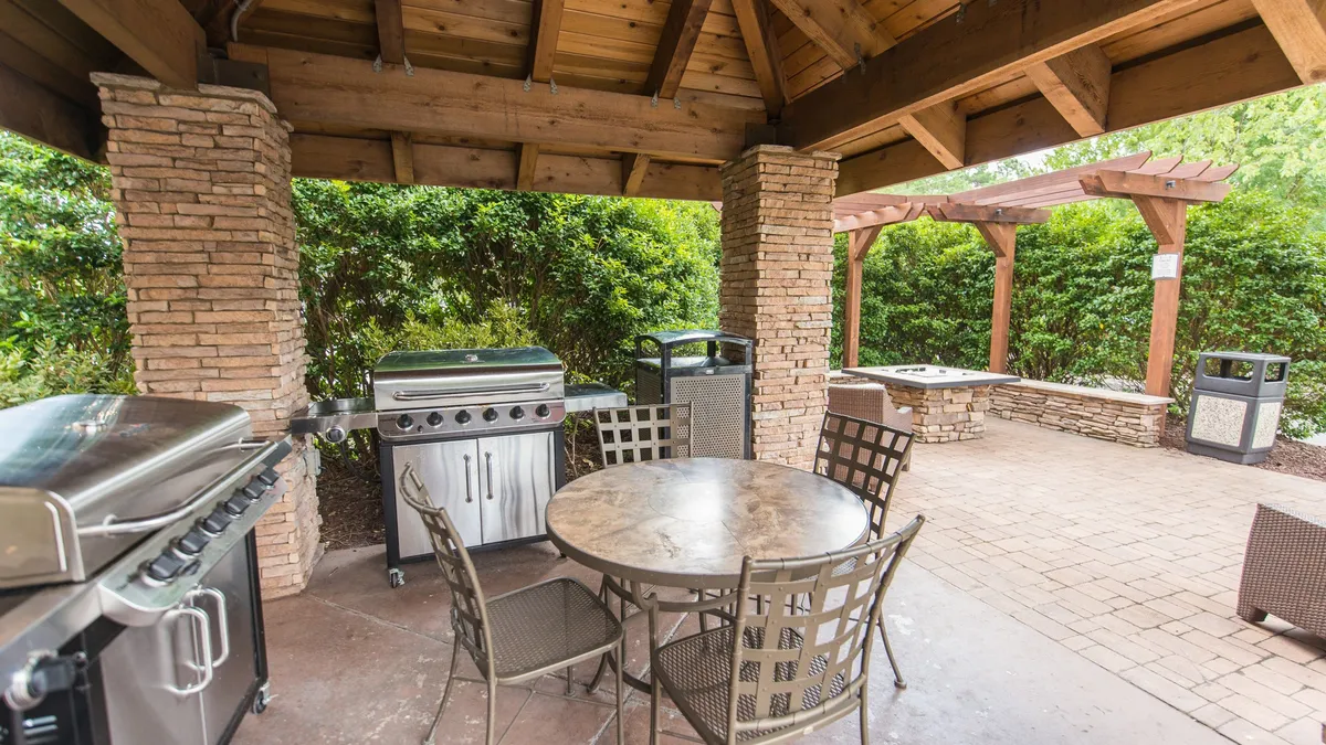 The image shows a well-equipped outdoor grilling station framed by stone pillars and surrounded by greenery. A spacious dining table invites families to gather for meals, while the relaxed mood is enhanced by the sunset light. The scene suggests opportunities for outdoor dining, celebrations, and togetherness.