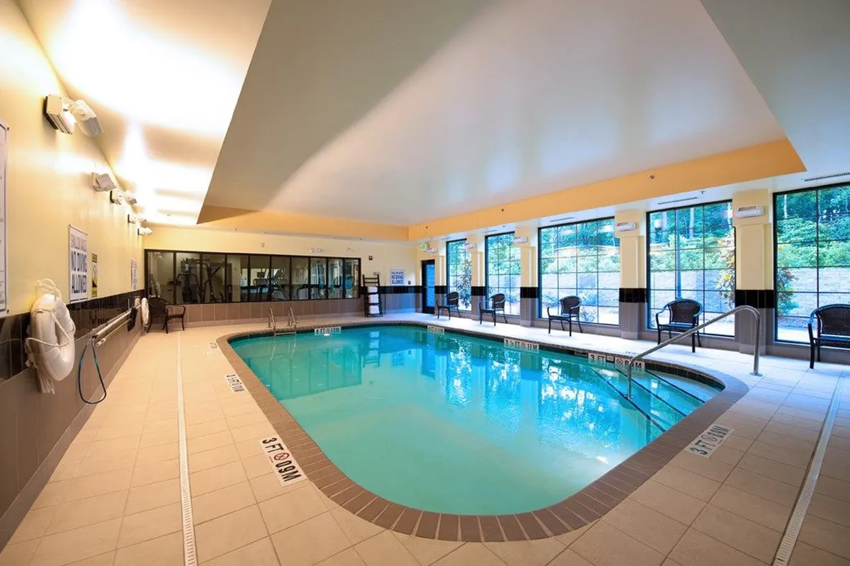 Bright and spacious indoor swimming pool area with large windows allowing natural light to flood the space. Comfortable lounge chairs line the pool, offering a relaxing environment for guests to unwind, socialize, or enjoy a quiet moment after work.
