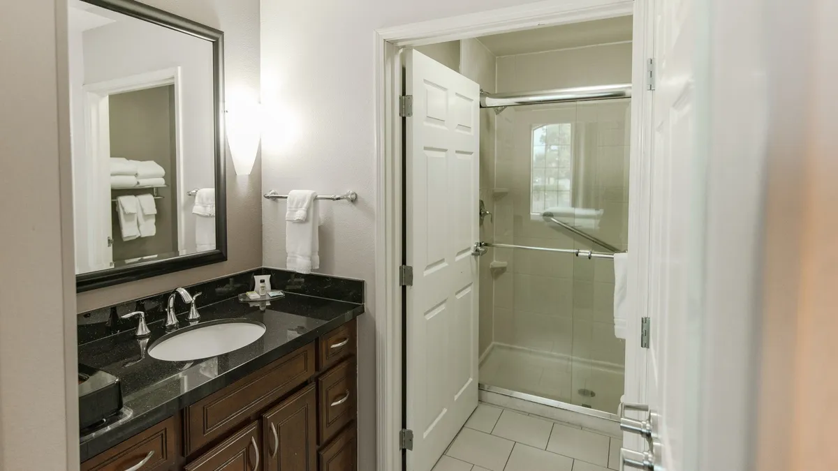 A contemporary bathroom featuring a spacious vanity with a sink, a large mirror, and a modern shower area. The space is bright and clean, evoking a sense of calm and refreshment—ideal for morning and evening routines. The design hints at both style and accessibility, supporting the description of spa-inspired baths.