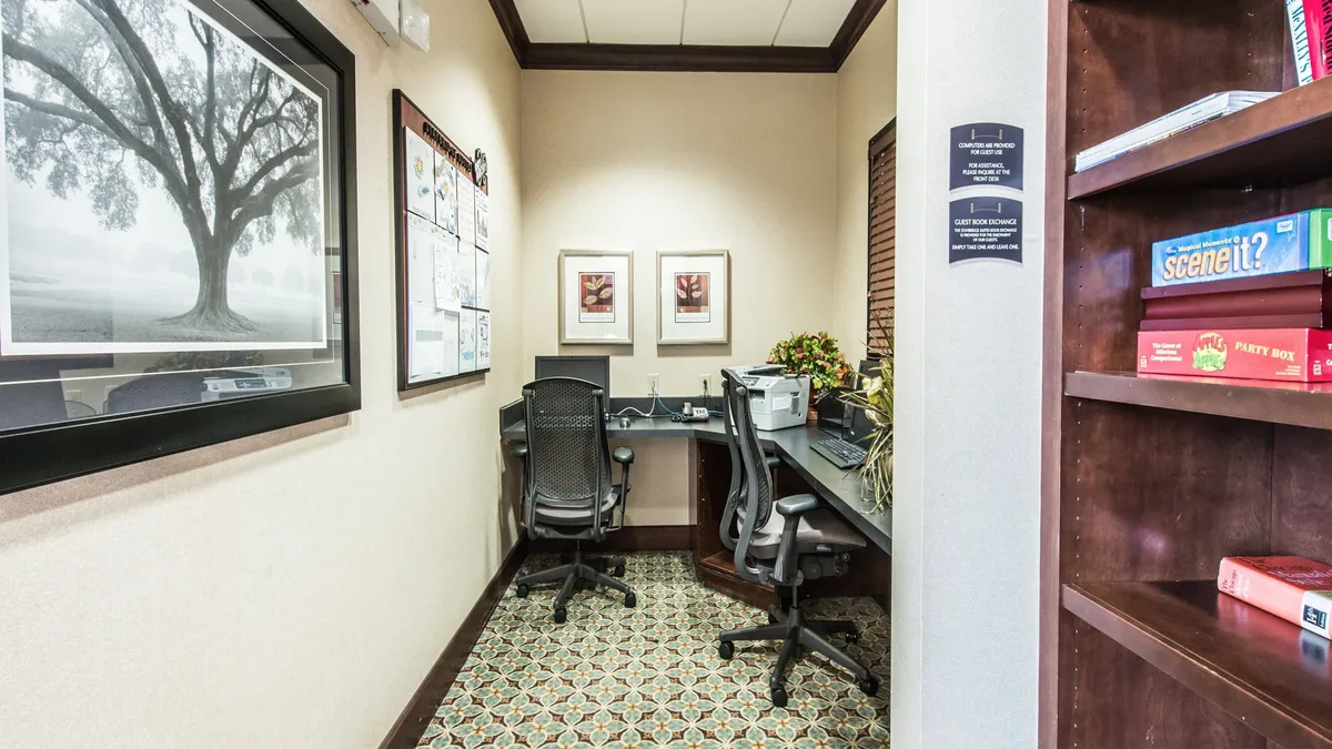 A thoughtfully designed business center space with two office desks, ergonomic chairs, a bookshelf filled with games and books, a large framed photograph on the wall, and a decorative plant, creating a productive yet welcoming atmosphere.