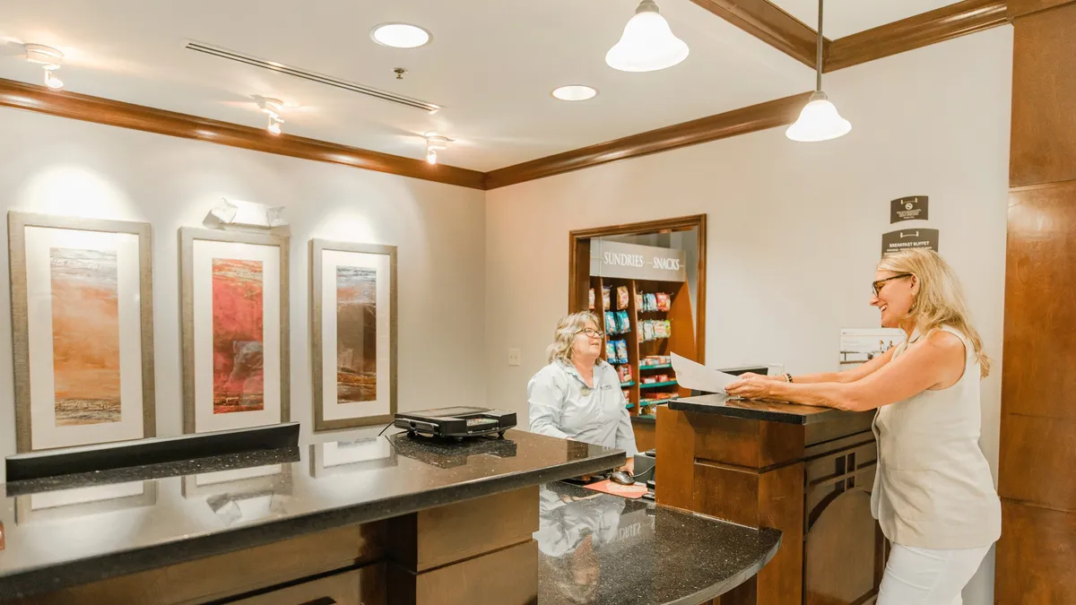 The image shows a welcoming reception area with a modern desk, stylish artwork on the wall, and a staff member engaging warmly with guests. The atmosphere is professional yet approachable, giving the impression of efficient service tailored to travelers' needs.