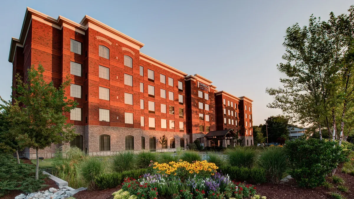 This image captures the exterior of the property, featuring a classic brick facade, a welcoming entrance awning, and vibrant flower beds. The scene is set during sunset, with the warm light enhancing the inviting atmosphere. The garden areas in the foreground hint at ample outdoor space for relaxation and play.